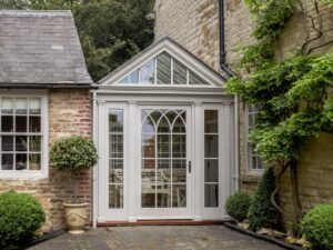A white double door with arched glass and a gabled roof, flanked by manicured shrubs and potted plants, leads into a cozy courtyard.