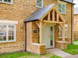  Charming stone house entrance with a wooden overhang and glass door, surrounded by well-maintained green lawns.